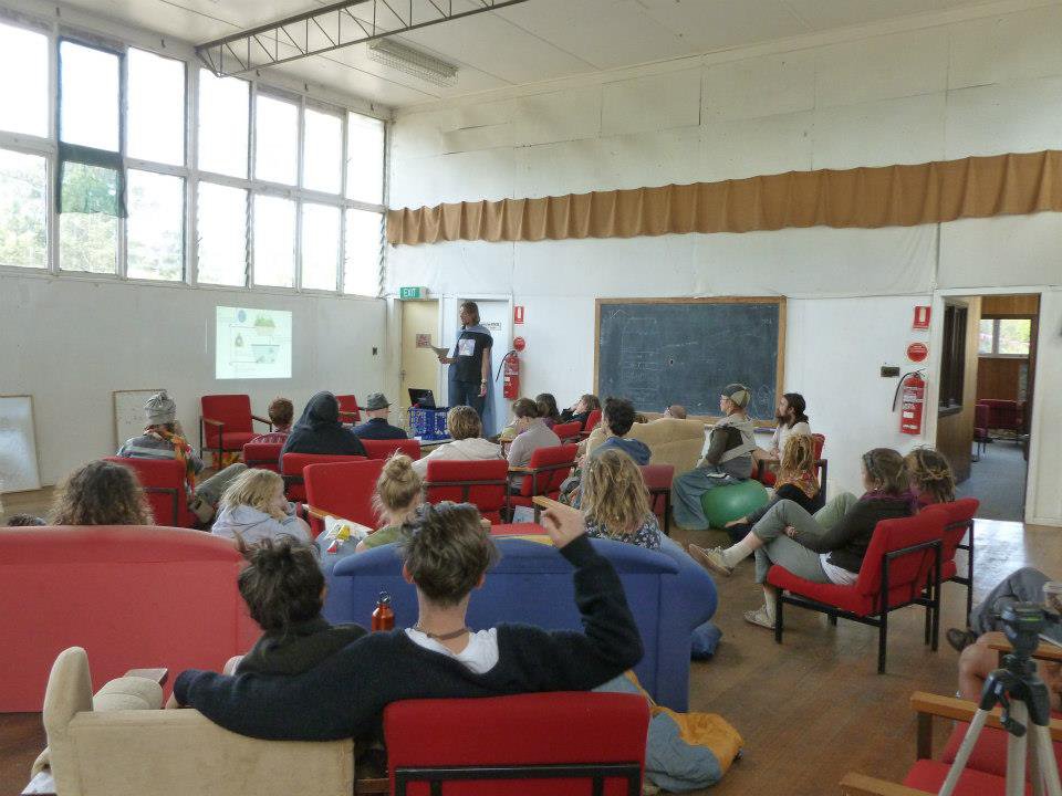 large high roofed room with presenter in corner using a data projector and about 20 people in shot listing