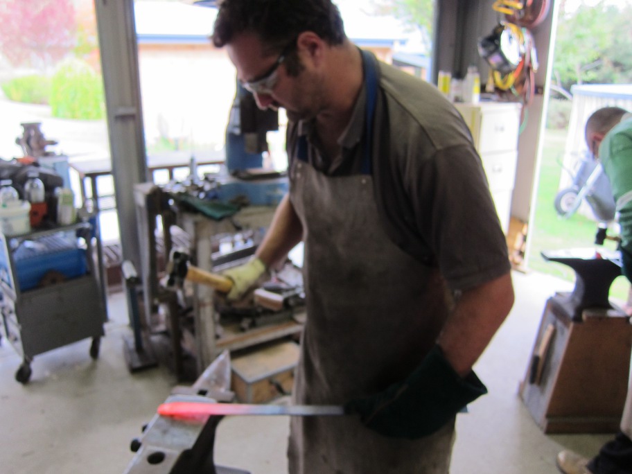 Adrian hammering a orange hot bar of metal into a knife on an anvil.