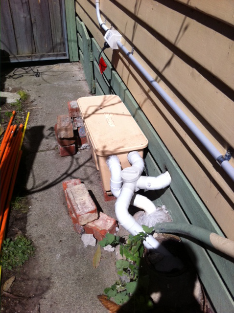 beige box situated on side of house low to ground, showing pip from shower and two way diversion to sewer or greywater box. Bricks protect the box from damage as it sits out into the path down the side of the house